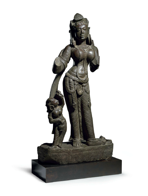 Lot 1608. A RARE AND IMPORTANT STONE FIGURE OF A MOTHER GODDESS  NORTH INDIA, ALMORA, 9TH CENTURY  This exquisitely carved and finely detailed sculpture depicts a goddess with her attendant. She is an idealized beauty with lotus-shaped eyes framed by delicately arched brows and centered by an incised spiral. Her lips are full and bow-shaped. Her finely incised hair is arranged in a simple twist above her left shoulder, with small curls escaping at her temples. She wears a tiara centered by a foliate element and with pendants terminating in floral buds and peepul leaves, both of which are echoed in her jewelry elsewhere. Her ample curves are highlighted by the multiple necklaces swaying over her breasts and belly, and a pendant girdle that encircles her hips. She stands with her weight on her right leg and her left turned out, causing her hip to sway to the right. She wears a long striated dhoti incised with flowers. A sash with floral motifs still encircles her upper right arm and shoulders. The attendant wears her hair in an identical manner and is clad in a dhoti and scarf with corresponding motifs. She holds a water pot and what appears to be a flower bud, flywhisk or small club in her raised hand. 26¾ in. (68 cms.) high  Estimate: $800,000-1,000,000. Provenance An important and distinguished private collection, Switzerland, before 1985. 